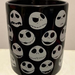 Puede incluir: Vaso de cer&aacute;mica negra con un patr&oacute;n repetido de caras blancas de Jack Skellington, cada una con una expresi&oacute;n diferente. El vaso cil&iacute;ndrico tiene un acabado brillante. El dise&ntilde;o est&aacute; inspirado en la pel&iacute;cula "Pesadilla antes de Navidad".