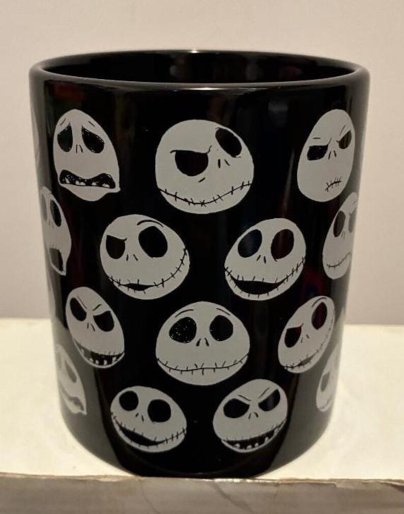 Puede incluir: Vaso de cer&aacute;mica negra con un patr&oacute;n repetido de caras blancas de Jack Skellington, cada una con una expresi&oacute;n diferente. El vaso cil&iacute;ndrico tiene un acabado brillante. El dise&ntilde;o est&aacute; inspirado en la pel&iacute;cula "Pesadilla antes de Navidad".
