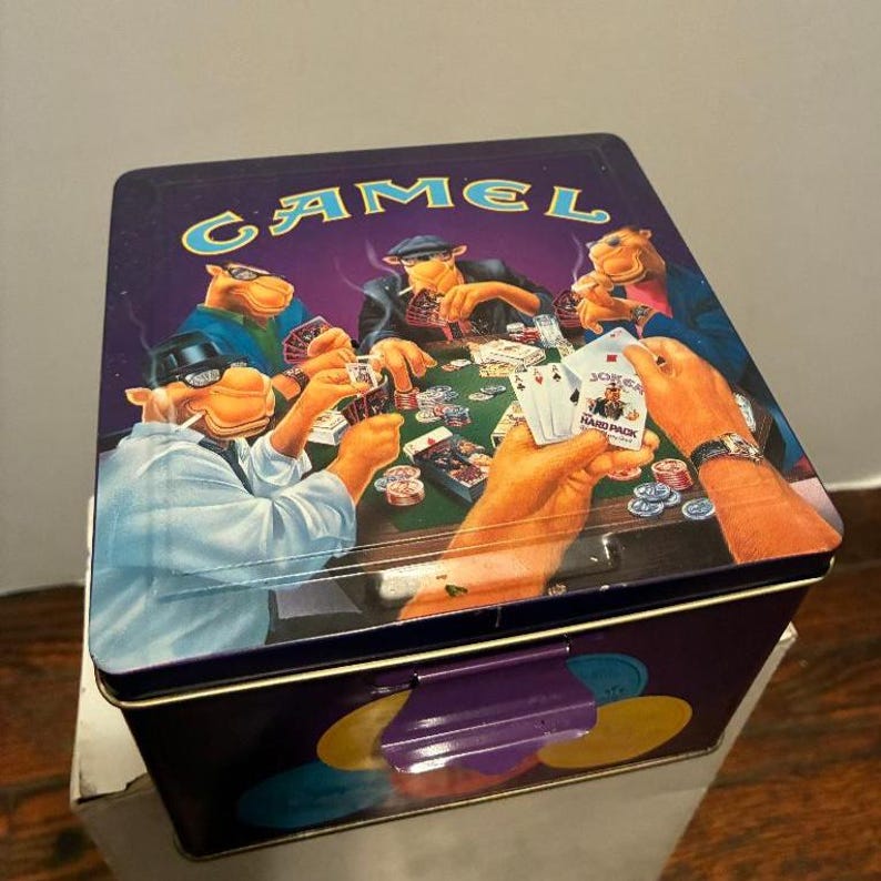 May include: A rectangular metal Camel-branded tin with a purple lid. The lid features a poker scene with cartoon camels playing cards. The tin has a purple handle and a design of colorful circles on the side.