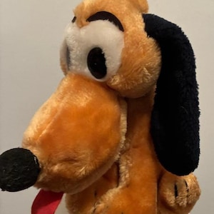 May include: A plush toy of Pluto, a cartoon dog. The toy has orange fur, black ears, a black nose, and a red tongue. The eyes are white with black pupils. The toy is a classic Disney character.