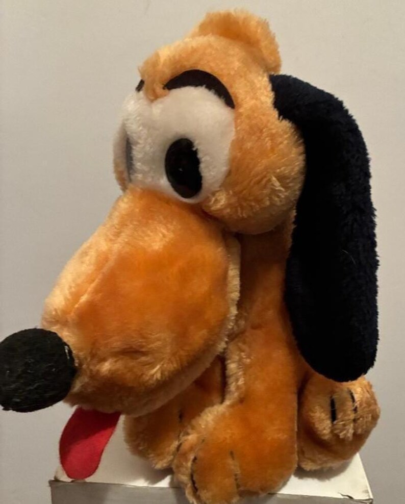 May include: A plush toy of Pluto, a cartoon dog. The toy has orange fur, black ears, a black nose, and a red tongue. The eyes are white with black pupils. The toy is a classic Disney character.
