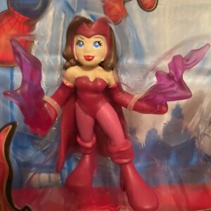 May include: A Scarlet Witch action figure in a dynamic pose, clad in a red and pink costume with matching boots and gloves. The figure has brown hair, blue eyes, and is holding translucent purple energy effects. The figure is in a clear plastic package.