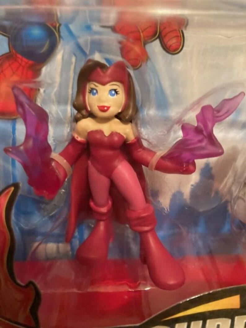 May include: A Scarlet Witch action figure in a dynamic pose, clad in a red and pink costume with matching boots and gloves. The figure has brown hair, blue eyes, and is holding translucent purple energy effects. The figure is in a clear plastic package.