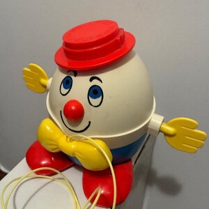 May include: A vintage, plastic toy shaped like a clown. The toy has a red hat, nose, and feet, with a yellow bow tie and hands. The clown's face is white with blue eyes and a smiling mouth. A yellow string is attached.