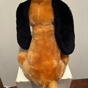 May include: A plush dog toy, viewed from the back, with a tan body and black floppy ears. The dog is sitting on a white surface. The tail is short and fluffy.