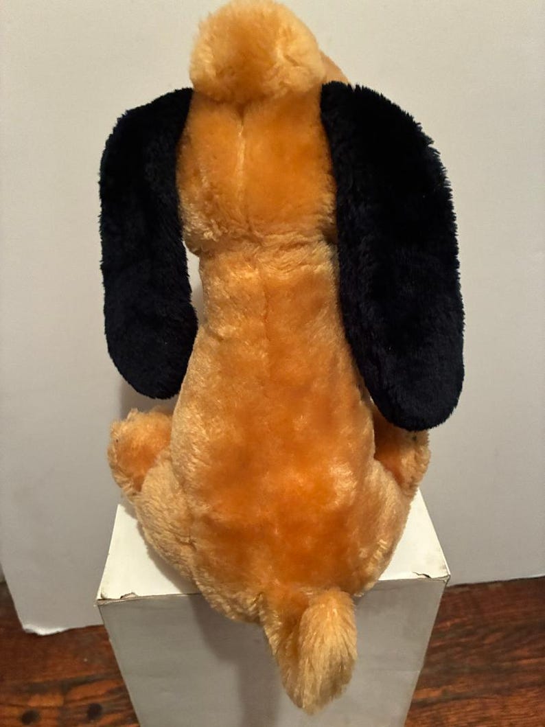 May include: A plush dog toy, viewed from the back, with a tan body and black floppy ears. The dog is sitting on a white surface. The tail is short and fluffy.