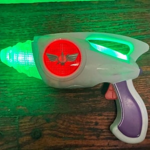 May include: A toy ray gun in shades of white, purple, and red, with a green light-up barrel and a red emblem. The handle is purple, and the trigger is red. The gun is designed to resemble a space blaster.