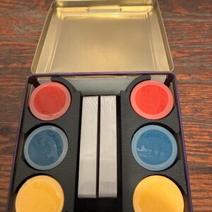 May include: An open, rectangular tin with a gold interior, holding a set of game chips and playing cards. The chips are red, blue, and yellow. The blue chips have a design and the word "CAMEL" printed on them. The cards are silver.