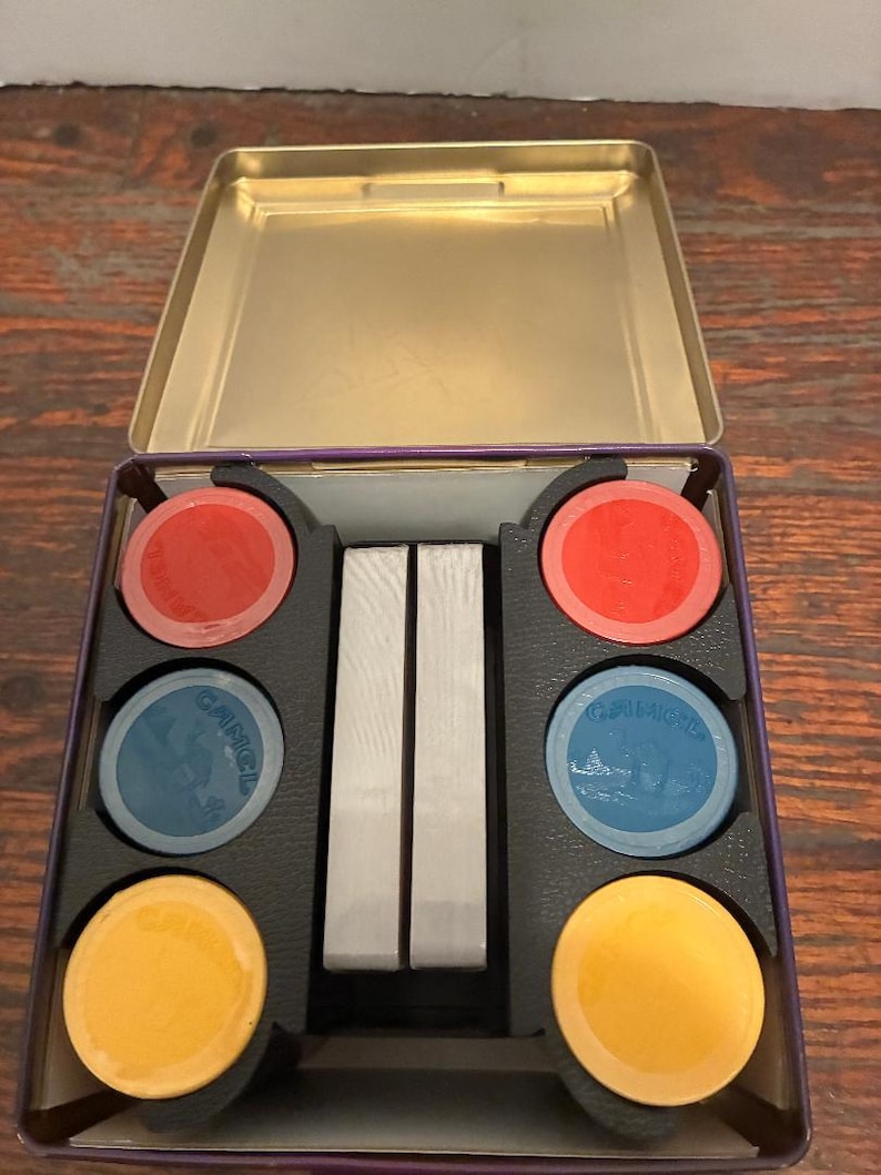 May include: An open, rectangular tin with a gold interior, holding a set of game chips and playing cards. The chips are red, blue, and yellow. The blue chips have a design and the word "CAMEL" printed on them. The cards are silver.