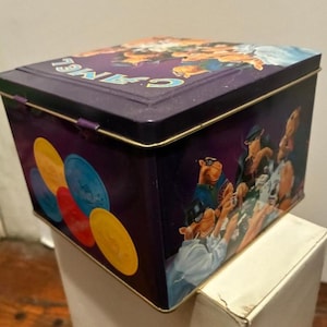 May include: A rectangular, purple metal tin with a hinged lid. The tin features a graphic of cartoon camels playing poker, with colorful poker chips on the side. The word "CAMEL" is printed on the lid in light blue.