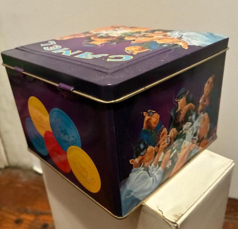 May include: A rectangular, purple metal tin with a hinged lid. The tin features a graphic of cartoon camels playing poker, with colorful poker chips on the side. The word "CAMEL" is printed on the lid in light blue.