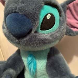May include: A plush toy character with dark blue fur, a large rounded nose, and oversized ears featuring pink inner linings. The character has light blue fur on the chest and around the eyes, with black and white embroidered eyes and small claws.