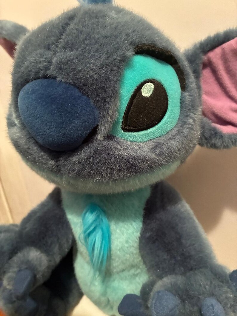May include: A plush toy character with dark blue fur, a large rounded nose, and oversized ears featuring pink inner linings. The character has light blue fur on the chest and around the eyes, with black and white embroidered eyes and small claws.