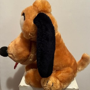 May include: A plush toy dog in a sitting position. The dog has orange fur, black ears, a black nose, and a red tongue sticking out. The toy is sitting on a white surface.