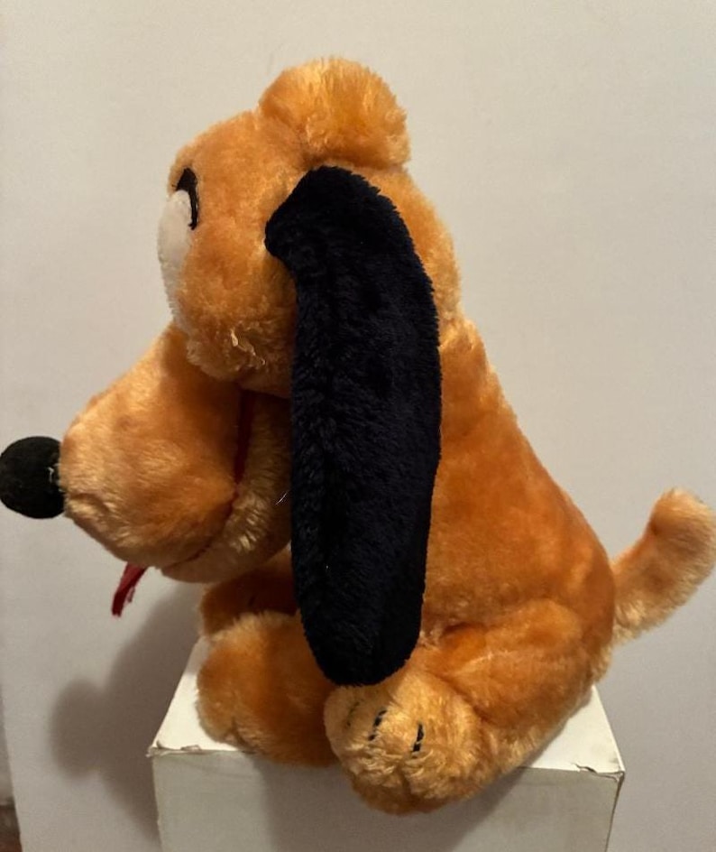 May include: A plush toy dog in a sitting position. The dog has orange fur, black ears, a black nose, and a red tongue sticking out. The toy is sitting on a white surface.