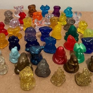 Puede incluir: Una colección de pequeñas figuras de colores en varias formas y colores, incluyendo rojo, azul, amarillo y verde. Algunas figuras son transparentes o brillantes. Las figuras están dispuestas sobre una superficie marrón claro.