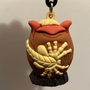 May include: A brown, red, and cream-colored keychain featuring a stylized, cartoonish design. The keychain has a round body with a cream-colored, leaf-like design on the front. The top is red, and the base is black.