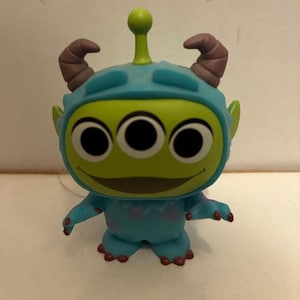 May include: A green alien figurine with three black eyes, wearing a blue helmet with brown horns. The alien has small arms and feet with red accents. The figurine is a collectible toy.