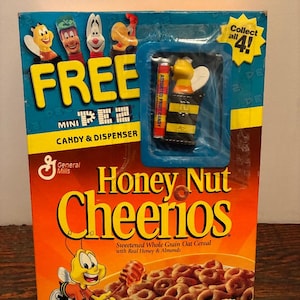 Könnte beinhalten: Eine Honey Nut Cheerios Müslipackung mit einem kostenlosen Mini-Pez-Bonbonspender. Die Schachtel ist orange und gelb mit einer Cartoon-Biene. Der Pez-Spender hat ein Bienen-Design. Auf der Schachtel steht auch "Sammle alle 4!"