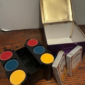 May include: A vintage game set featuring a black chip holder with red, blue, and yellow chips. The set includes two decks of Camel brand playing cards and a purple and gold metal box.
