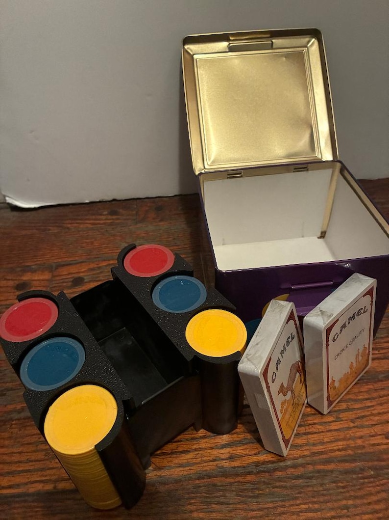 May include: A vintage game set featuring a black chip holder with red, blue, and yellow chips. The set includes two decks of Camel brand playing cards and a purple and gold metal box.