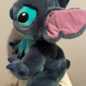 May include: A plush toy of the character Stitch with dark blue fur, large ears with light pink inner lining, and bright turquoise accents on the chest and around the eyes. The figure features a tuft of blue hair on top and dark blue claws.