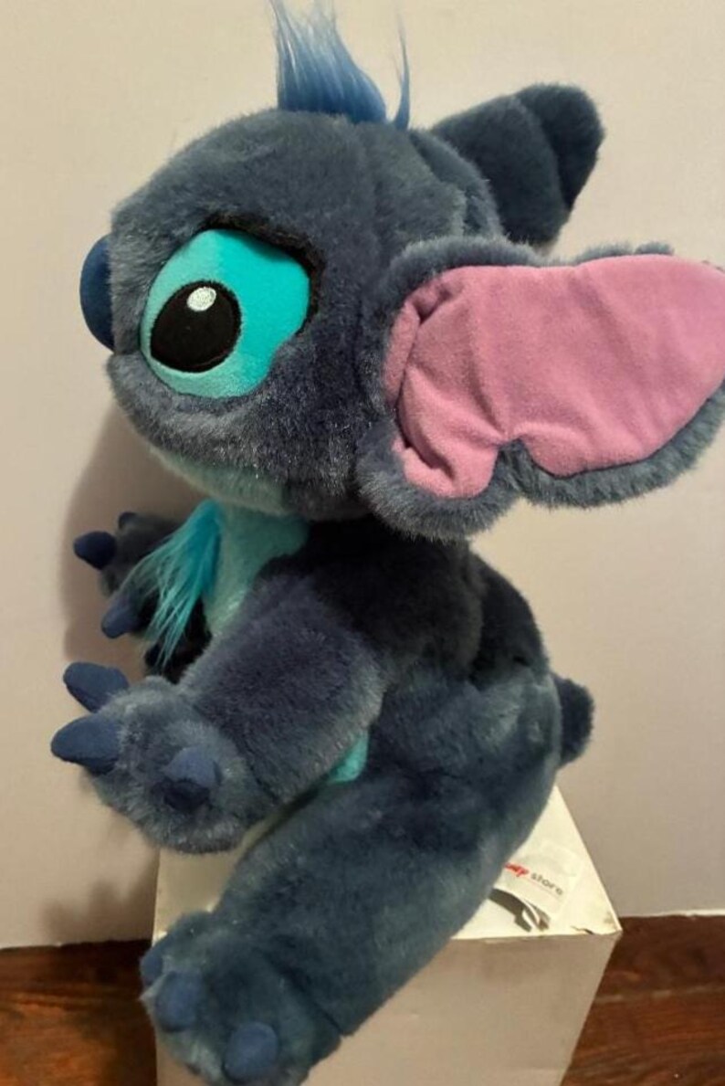 May include: A plush toy of the character Stitch with dark blue fur, large ears with light pink inner lining, and bright turquoise accents on the chest and around the eyes. The figure features a tuft of blue hair on top and dark blue claws.