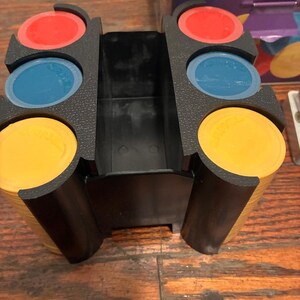 May include: A black plastic game chip holder with stacks of yellow, red, and blue chips. The holder has three slots, each holding a stack of chips. The chips are round and have the word "SPADES" printed on them.