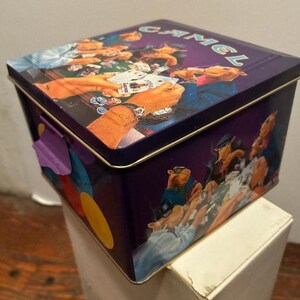 May include: A rectangular purple metal tin with a hinged lid. The lid and sides feature a colorful illustration of camels playing poker. The word "CAMEL" is printed on the lid in a light blue color. The tin has a purple clasp.