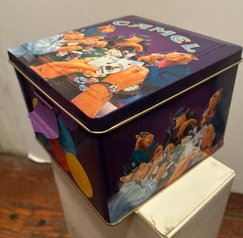 May include: A rectangular purple metal tin with a hinged lid. The lid and sides feature a colorful illustration of camels playing poker. The word "CAMEL" is printed on the lid in a light blue color. The tin has a purple clasp.