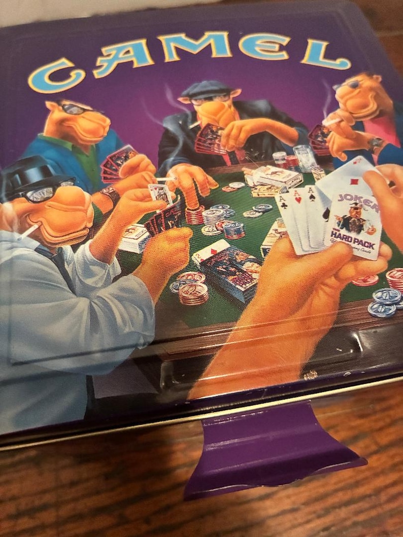 May include: A vintage Camel cigarette tin featuring a poker game scene with anthropomorphic camels. The tin is primarily purple with the word "CAMEL" in large, yellow letters. The poker scene includes playing cards, chips, and the text "JOKER HARD PACK".