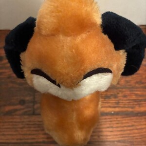 May include: A plush fox toy with orange fur, black ears, and a black nose. The fox has a white face and closed eyes. The toy is made of soft material and is designed to look like a fox.