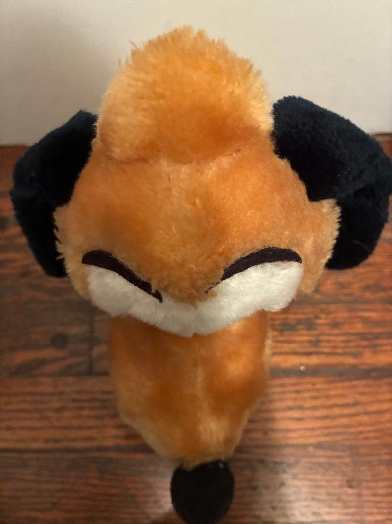 May include: A plush fox toy with orange fur, black ears, and a black nose. The fox has a white face and closed eyes. The toy is made of soft material and is designed to look like a fox.