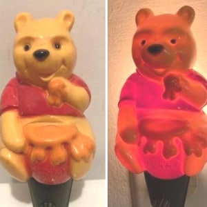 Może przedstawiać: Nocna lampka Winnie the Pooh w kolorze żółtym z czerwoną koszulą i bębnem. Lampka jest włączona i świeci na pomarańczowo.