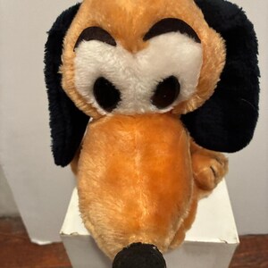 May include: A plush toy dog with a tan body, black ears, and a black nose. The dog has large, round black eyes and a white face. The toy is sitting on a white surface.