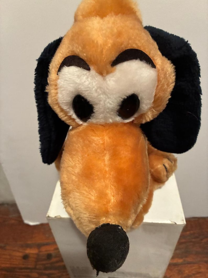 May include: A plush toy dog with a tan body, black ears, and a black nose. The dog has large, round black eyes and a white face. The toy is sitting on a white surface.