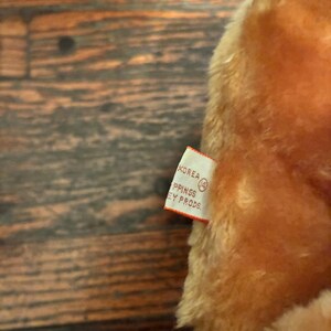 May include: Close-up of a plush toy with a tag. The toy is a warm, light brown color. The tag is white with black text that reads "KOREA" and "PPINGS EY PRODS."
