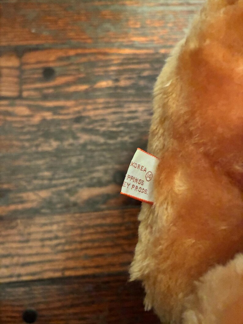 May include: Close-up of a plush toy with a tag. The toy is a warm, light brown color. The tag is white with black text that reads "KOREA" and "PPINGS EY PRODS."