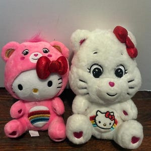 May include: Two plush toys: one pink, one white. The pink toy is a Hello Kitty character wearing a bear-like hood and a red bow. The white toy has a red bow and a Hello Kitty emblem on its chest. Both toys have embroidered details.