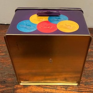 May include: A purple metal storage tin with a gold interior. The lid features colorful circular designs with camel illustrations. The tin has a rectangular shape with a hinged lid and a small handle.