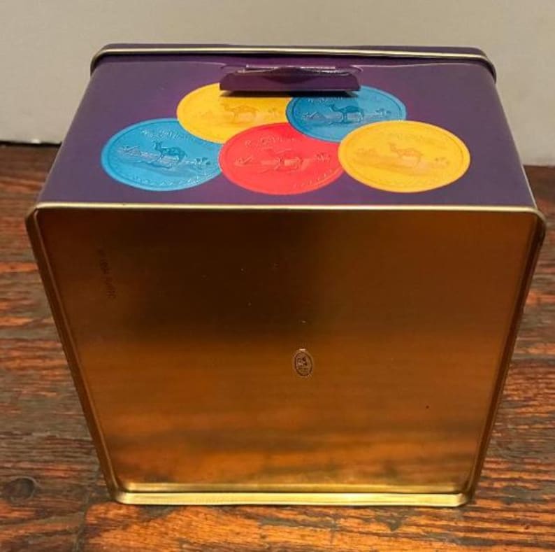May include: A purple metal storage tin with a gold interior. The lid features colorful circular designs with camel illustrations. The tin has a rectangular shape with a hinged lid and a small handle.