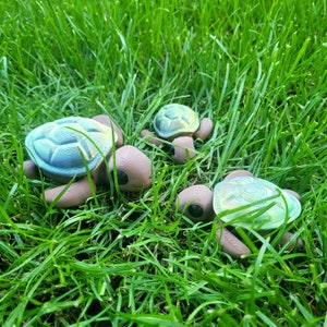 Könnte beinhalten: Drei dekorative Schildkrötenfiguren mit braunen Körpern und strukturierten, irisierenden blau-grünen Schalen liegen im grünen Gras. Die Schildkröten sind unterschiedlich groß, die größte im Vordergrund.