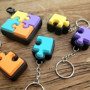 Könnte beinhalten: Vier farbige Puzzle-Schlüsselanhänger mit schwarzen Metallringen. Die Schlüsselanhänger sind aus Kunststoff und haben verschiedene Farben, darunter Blau, Gelb, Lila und Orange.