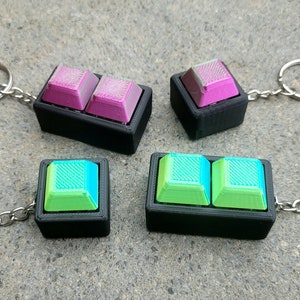 Könnte beinhalten: Vier Schlüsselanhänger mit Keycaps, zwei mit grünen und blauen Keycaps und zwei mit rosa und violetten Keycaps. Die Keycaps sind in schwarzen Plastikrahmen mit einer silbernen Kette befestigt.