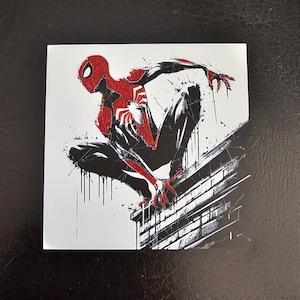 May include: A black and red superhero, Spider-Man, is perched on a brick wall. He is in a crouched position with his arms outstretched. The background is white with black splatters of paint.