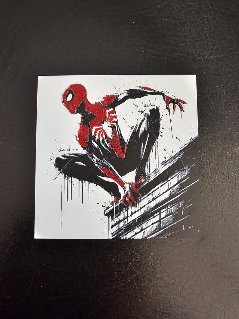 May include: A black and red superhero, Spider-Man, is perched on a brick wall. He is in a crouched position with his arms outstretched. The background is white with black splatters of paint.