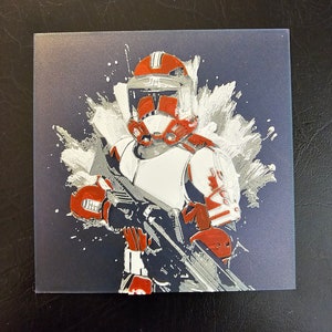 Può includere: Un'illustrazione dipinta di bianco e rosso di un clone trooper di Star Wars con un fucile blaster. Lo sfondo è blu scuro con schizzi bianchi.