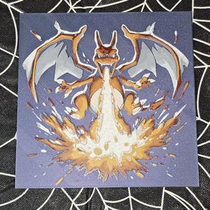 May include: Square canvas art featuring a Charizard illustration. The artwork depicts the fire-breathing dragon in shades of orange, white, and brown against a blue background. The dragon is shown breathing fire, with a burst of orange flames.