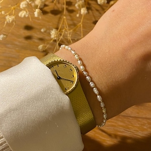 Könnte beinhalten: Eine goldfarbene Armbanduhr mit hellgrünem Armband wird mit einem zarten Perlenarmband kombiniert. Das Zifferblatt der Uhr zeigt schwarze Ziffern. Das Handgelenk ist mit dem Schmuck vor dem Hintergrund getrockneter Blumen geschmückt.
