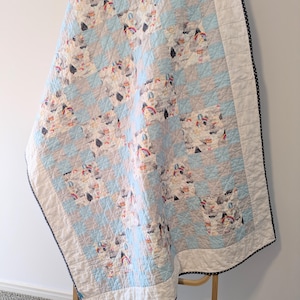 May include: A patchwork quilt featuring a pattern of hot air balloons, rainbows, and clouds in shades of light blue and grey. The quilt has a white border with a black and white polka dot trim. It is displayed over a wooden ladder.